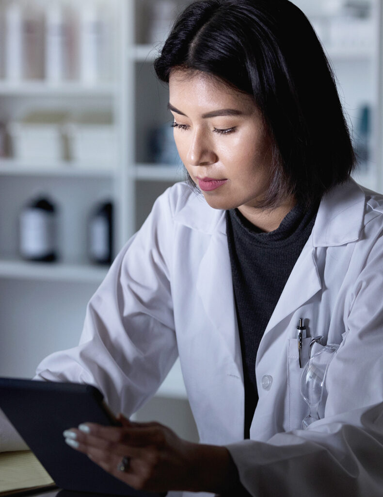 Female pharmacist in a lab coat reviewing information on a tablet, with pharmacy shelves and bottles in the background